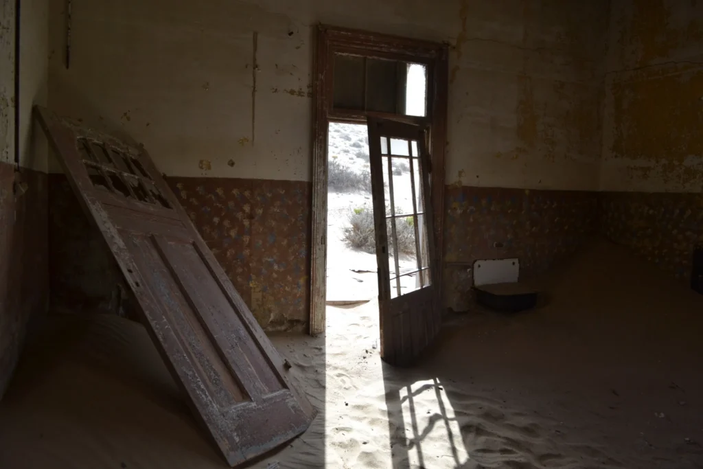 Interno buio abbandonato a Kolmanskop, con porte danneggiate e sabbia sul pavimento