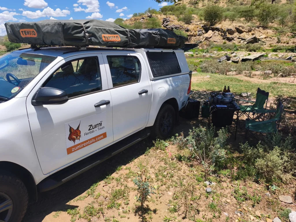 Toyota Hilux di Zumi con tende da tetto e attrezzatura da campeggio allestite accanto a un fiume in Namibia